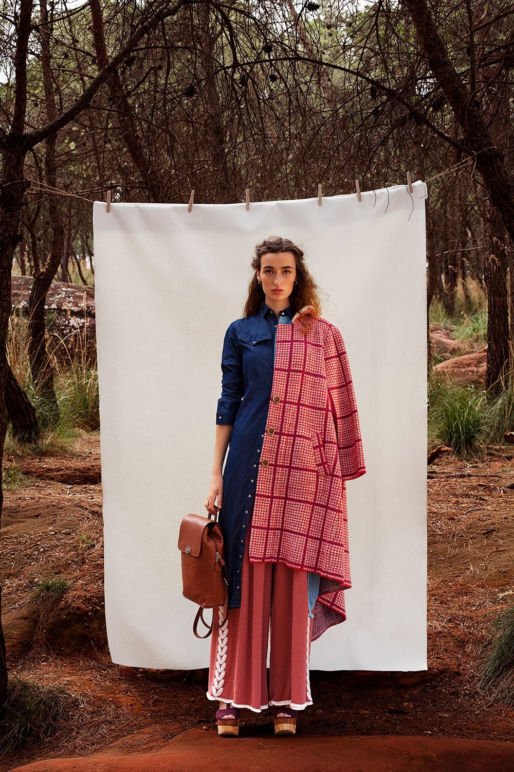 Model in a denim shirt dress and red houndstooth coat standing before a white sheet hung in a pine forest, tan leather backpack